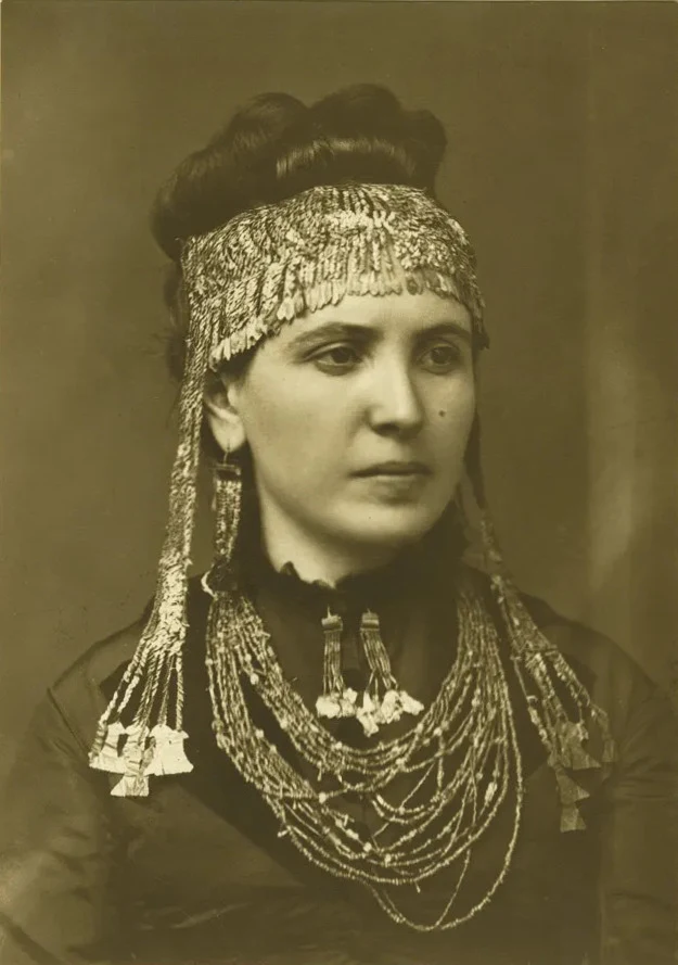 Sophia Schliemann ubrana w biżuterię ze zbiorów Skarbu króla Priama. Kobieta w tradycyjnym stroju, ozdobiona bogato zdobioną biżuterią na głowie i wieloma warstwami naszyjników z błyszczących łańcuszków, patrząca w bok na neutralnym tle.