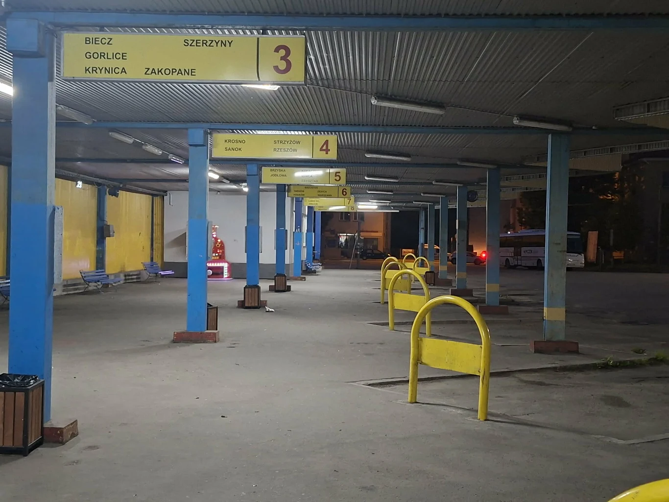 Puste stanowiska autobusowe oznaczone żółtymi tablicami z numerami i kierunkami, rozmieszczone pod metalowym zadaszeniem, z żółtymi barierkami oddzielającymi perony.