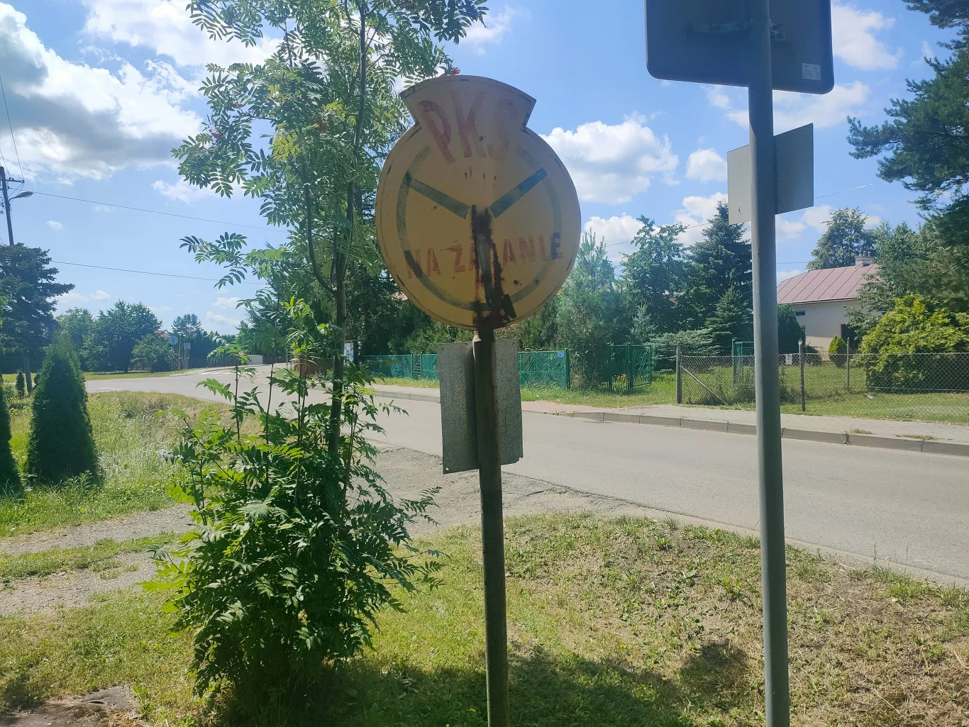 Zniszczony znak PKP w otoczeniu zieleni, przy bocznej asfaltowej drodze prowadzącej przez spokojną wiejską okolicę.