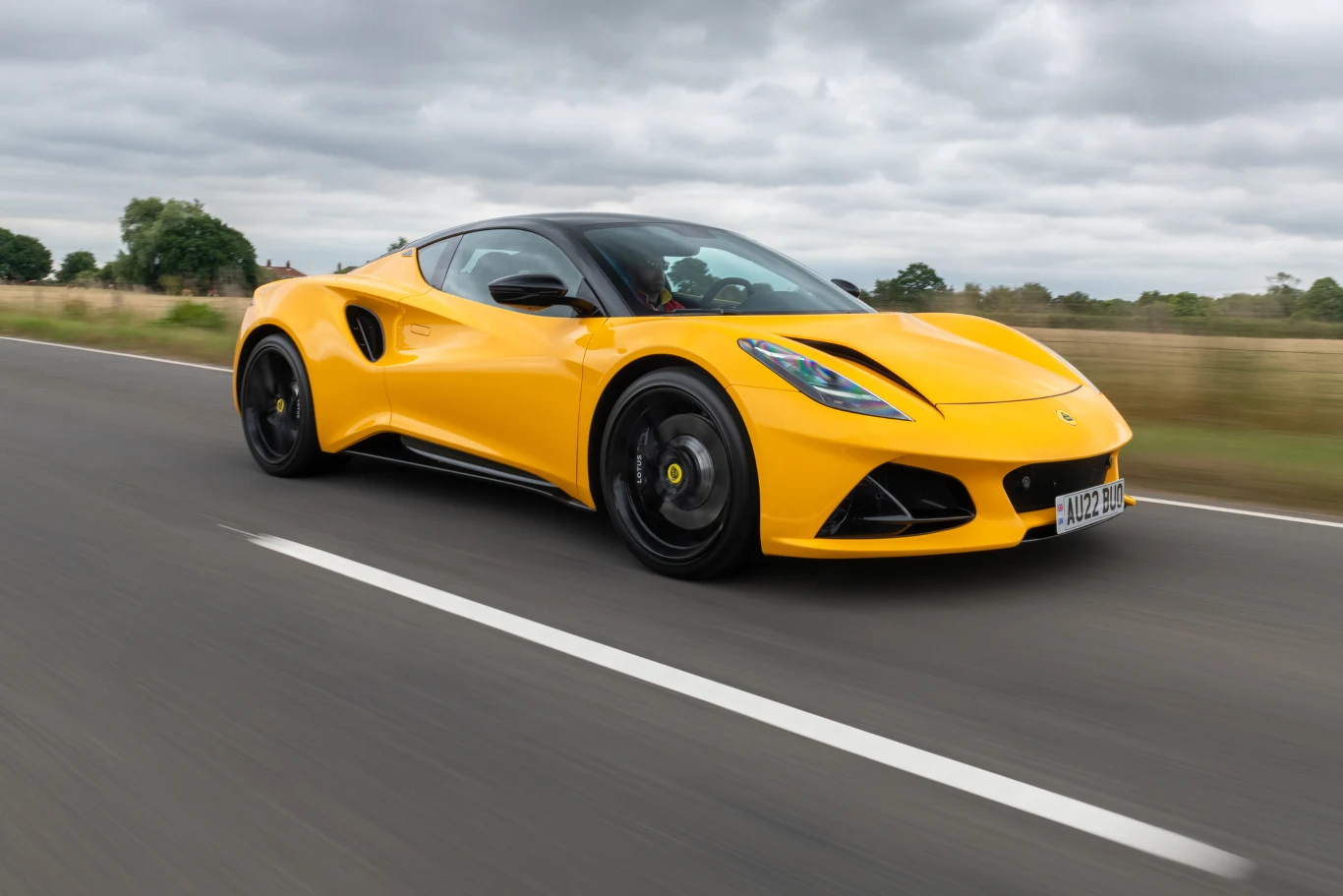 Lotus Emira już w Polsce. Ceny rozpoczynają się od 496 tys. zł. Jaskrawożółty Lotus Emira na pustej asfaltowej drodze, otoczonej polami i niską roślinnością