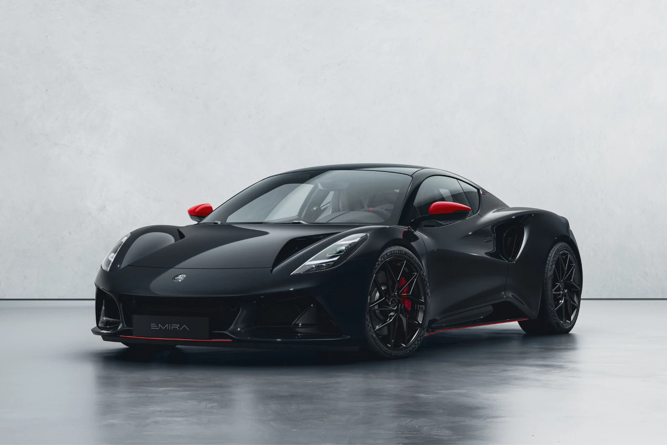 Lotus Emira Turbo SE. Czarny sportowy samochód z czerwonymi akcentami na lusterkach i felgach, stojący na jasnej, gładkiej powierzchni, wyróżniający się opływową sylwetką oraz agresywną linią przednich reflektorów.