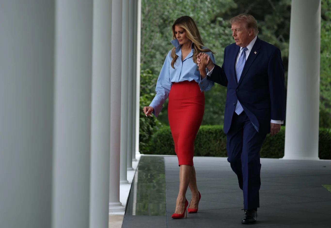 Uwagę także przyciągnęła stylizacja Melanii Elegancka kobieta w czerwonej spódnicy i niebieskiej koszuli idzie pod ramię z mężczyzną w granatowym garniturze wzdłuż kolumnady na zewnątrz budynku.