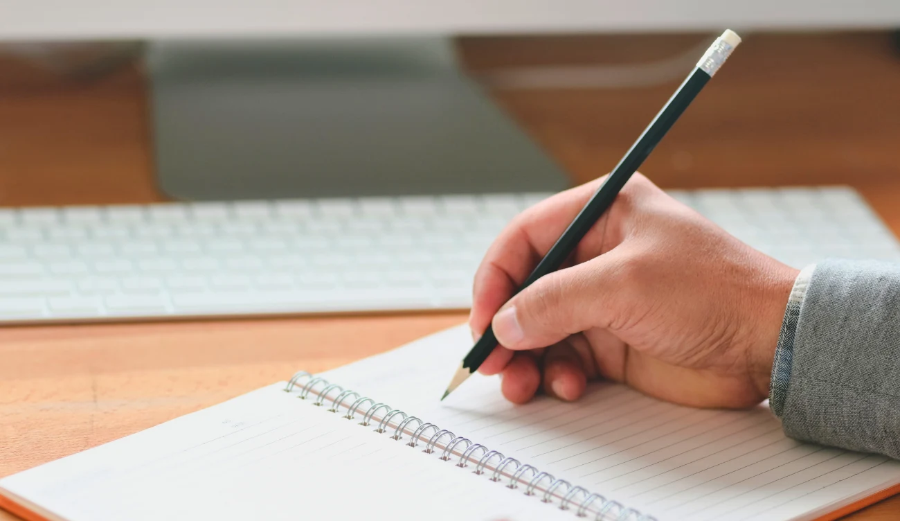 Zagadka matematyczna dla geniuszy? Ręka z ołówkiem pisząca w zeszycie na biurku, obok klawiatura komputerowa, temat: zagadka matematyczna.