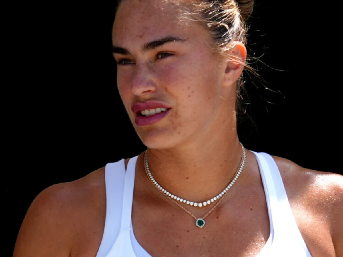 Aryna Sabalenka Kobieta w sportowym stroju, z upiętymi włosami i naszyjnikiem z zielonym kamieniem, widoczna na ciemnym tle.