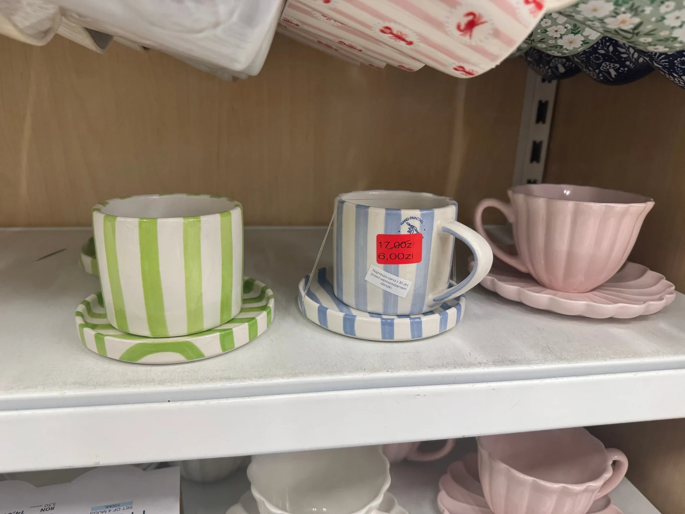 Kubki w Pepco Trzy filiżanki ze spodkami ustawione na półce, dwie w paski o barwach zielonych i niebieskich, jedna różowa w stylu retro, wszystkie z ceramiki i delikatnym wykończeniem.