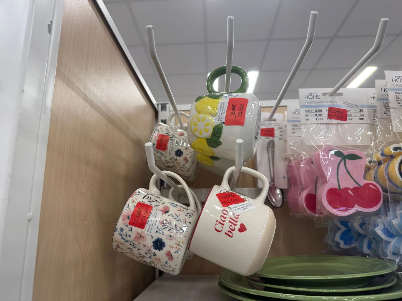 Kubki Kilka kolorowych kubków z motywami kwiatów, cytryn i napisem „Ciao bella” zawieszonych na metalowych haczykach, obok tekturowych zawieszek z cenami oraz dekoracyjnych magnesów w kształcie wiśni.