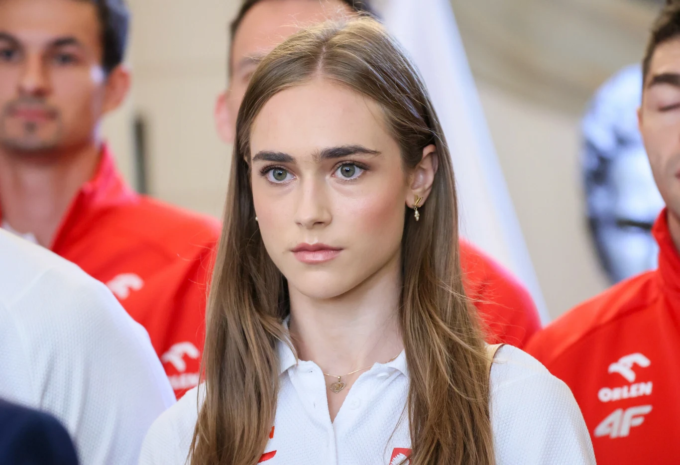 Młoda kobieta o długich, prostych włosach ubrana w białą koszulkę sportową z logo 4F, stojąca na pierwszym planie w otoczeniu osób w czerwonych strojach z tym samym znakiem firmowym.