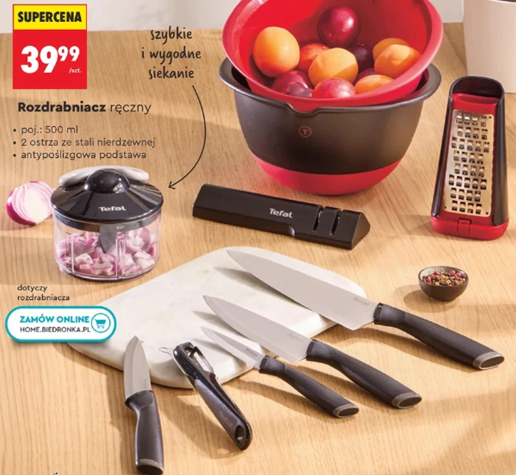 Kuchenne przybory Tefal od 39,99 zł Deska do krojenia z kompletem czterech noży i obieraczką, obok ręczny rozdrabniacz z posiekaną cebulą, w tle duża miska z owocami, tarka oraz młynek do przypraw.
