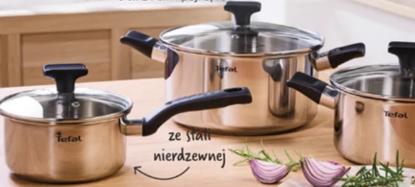 Zestaw 2 garnków Tefal za 229 zł Trzy garnki marki Tefal ze stali nierdzewnej z czarnymi uchwytami i szklanymi pokrywkami ustawione na drewnianym blacie, obok połówki czerwonej cebuli i gałązki rozmarynu.