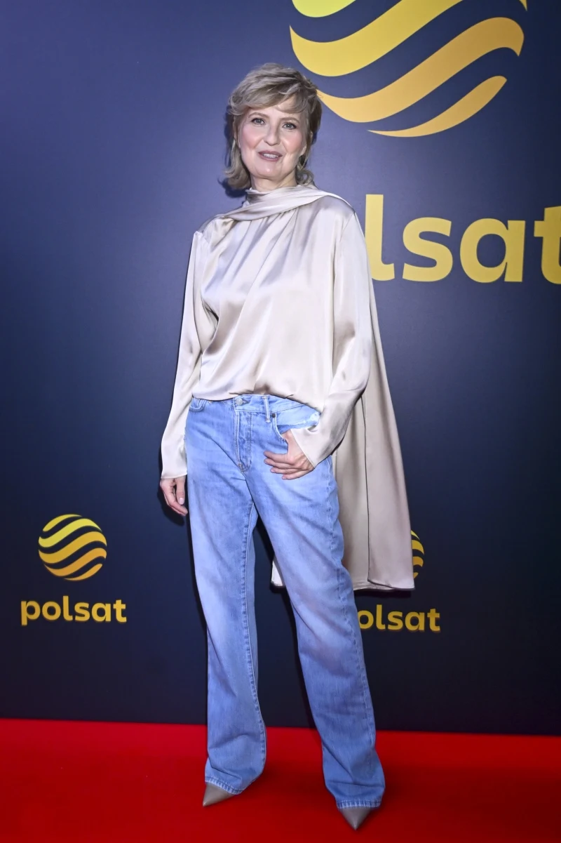 Kobieta w jasnej, luźnej bluzce z połyskiem i szerokich, niebieskich jeansach, stojąca na czerwonym dywanie na tle ścianki z logo Polsat.