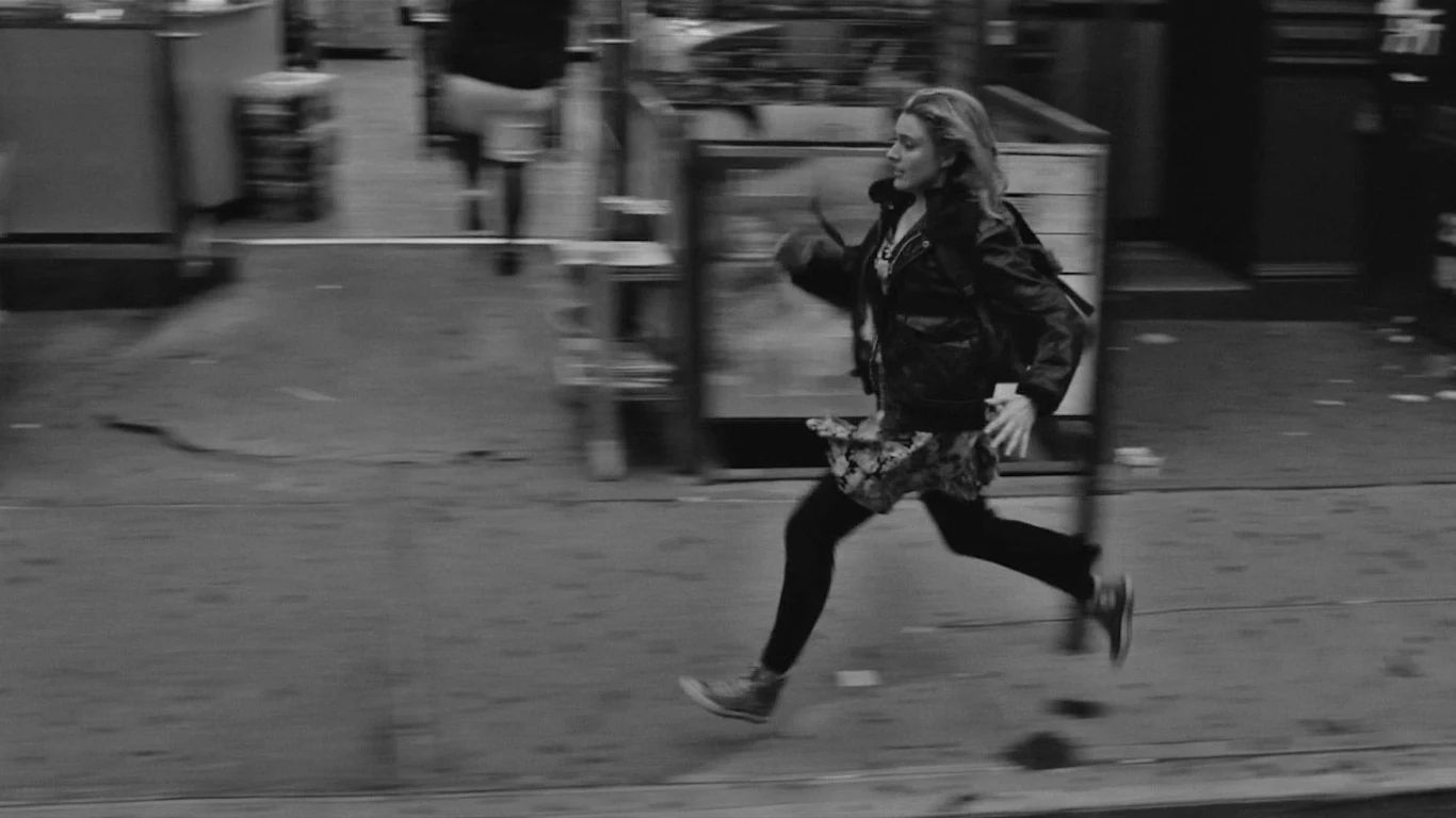 Greta Gerwig w scenie z filmu "Frances Ha" Greta Gerwig biegnie chodnikiem miejskiej ulicy w kurtce i sukience w scenie z filmu "Frances Ha".