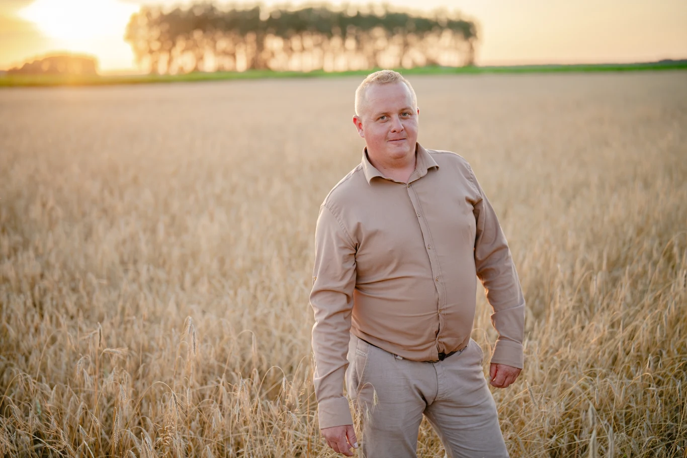 Mężczyzna w beżowej koszuli i spodniach stoi na środku pola zboża podczas złotej godziny, w tle widoczny szpaler drzew na horyzoncie.