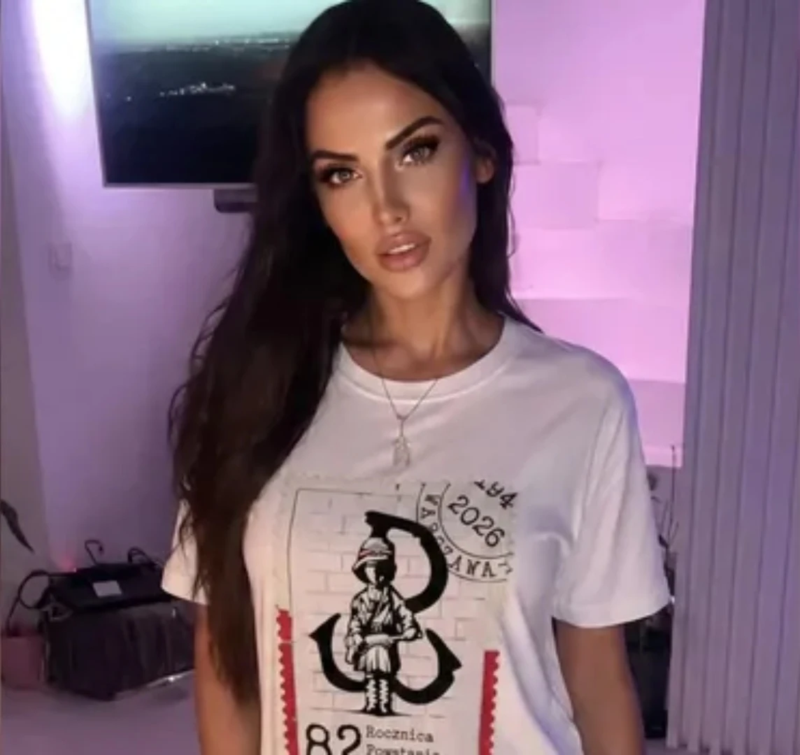 Monika Laskowska Młoda kobieta z długimi, ciemnymi włosami ubrana w białą koszulkę z nadrukiem powstańca warszawskiego, akcentowana delikatnym makijażem w minimalistycznym, nowoczesnym wnętrzu o fioletowym oświetleniu.