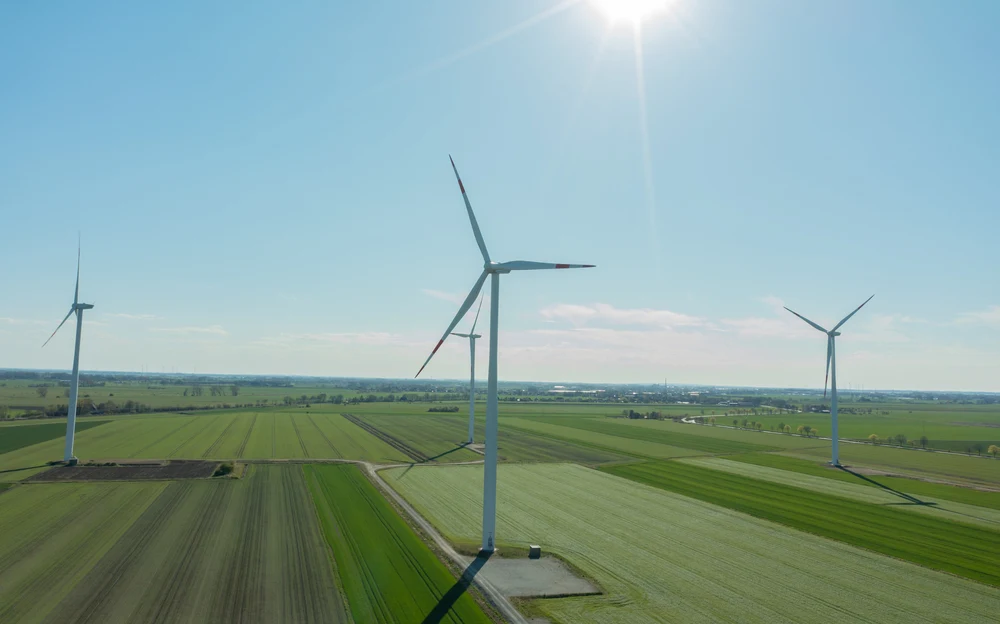PSEW Trzy wysokie turbiny wiatrowe rozmieszczone na zielonych polach uprawnych pod niebieskim, słonecznym niebem.