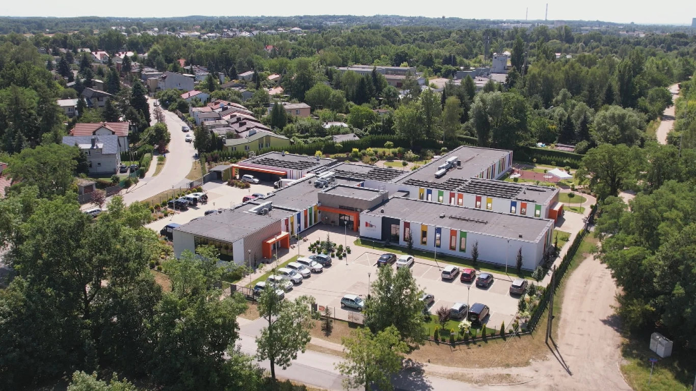 Nowoczesny kompleks budynków o jasnej elewacji z kolorowymi akcentami, otoczony ogrodzeniem i parkingiem, położony w zielonej dzielnicy z zabudową jednorodzinną.