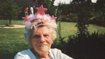 Starsza kobieta z siwymi włosami siedzi na krześle w ogrodzie, ma na głowie różową opaskę z gwiazdkami i napisem "Birthday Girl", uśmiecha się i nosi biały sweter oraz korale.