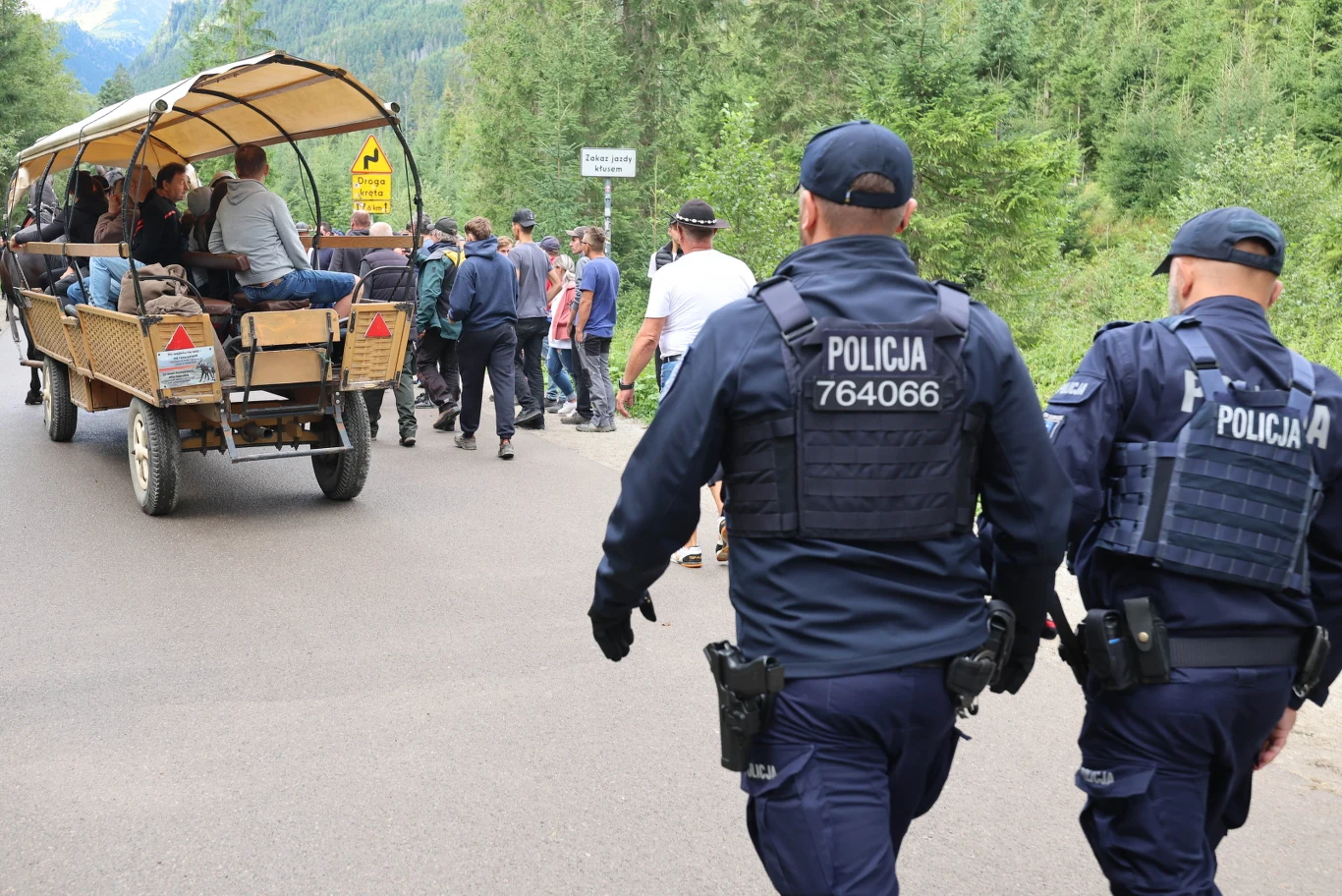 Dwóch policjantów w umundurowaniu patroluje drogę w lesie, obok której znajduje się grupa osób i zaprzęg konny z pasażerami siedzącymi na drewnianej platformie zadaszonej plandeką.