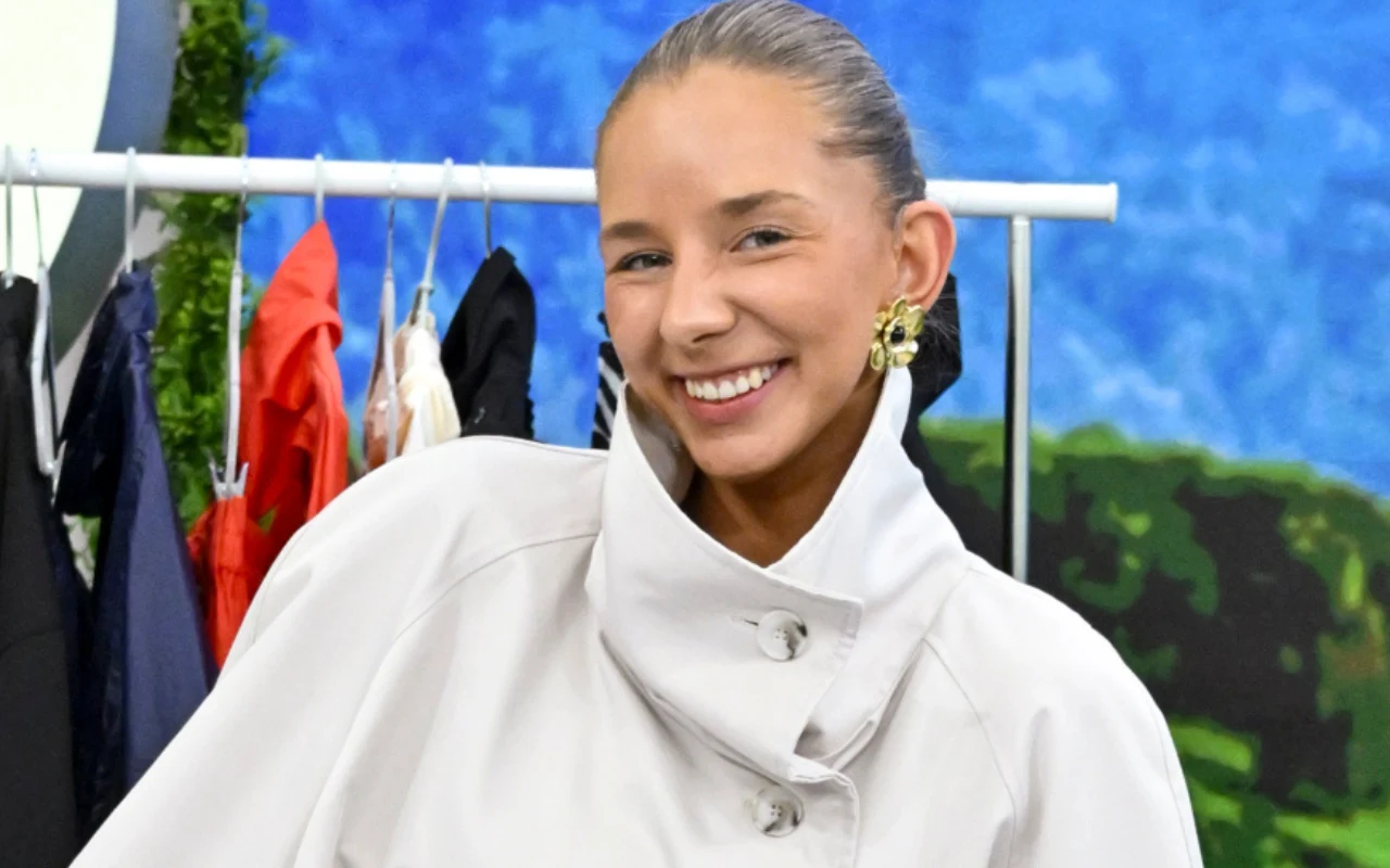 Stylistka Karolina Wolska Uśmiechnięta kobieta w jasnym płaszczu i oryginalnych złotych kolczykach na tle wiszących ubrań oraz niebiesko-zielonej dekoracji.