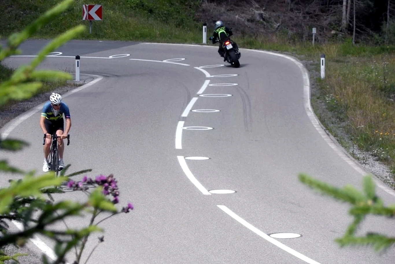 Okręgi mają zmieniać tor jazdy motocyklistów Kręta górska droga z białymi okręgami na jezdni. Motocyklista i rowerzysta na osobnych pasach.