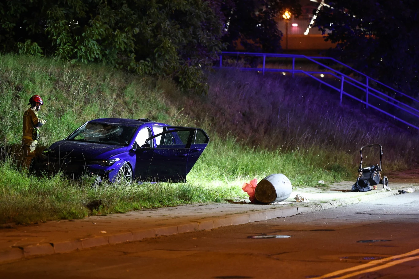 Tragiczny wypadek w Krakowie. Dwie osoby nie żyją Uszkodzony samochód osobowy przy trawie, otwarte drzwi, przewrócony wózek dziecięcy i strażak w hełmie.