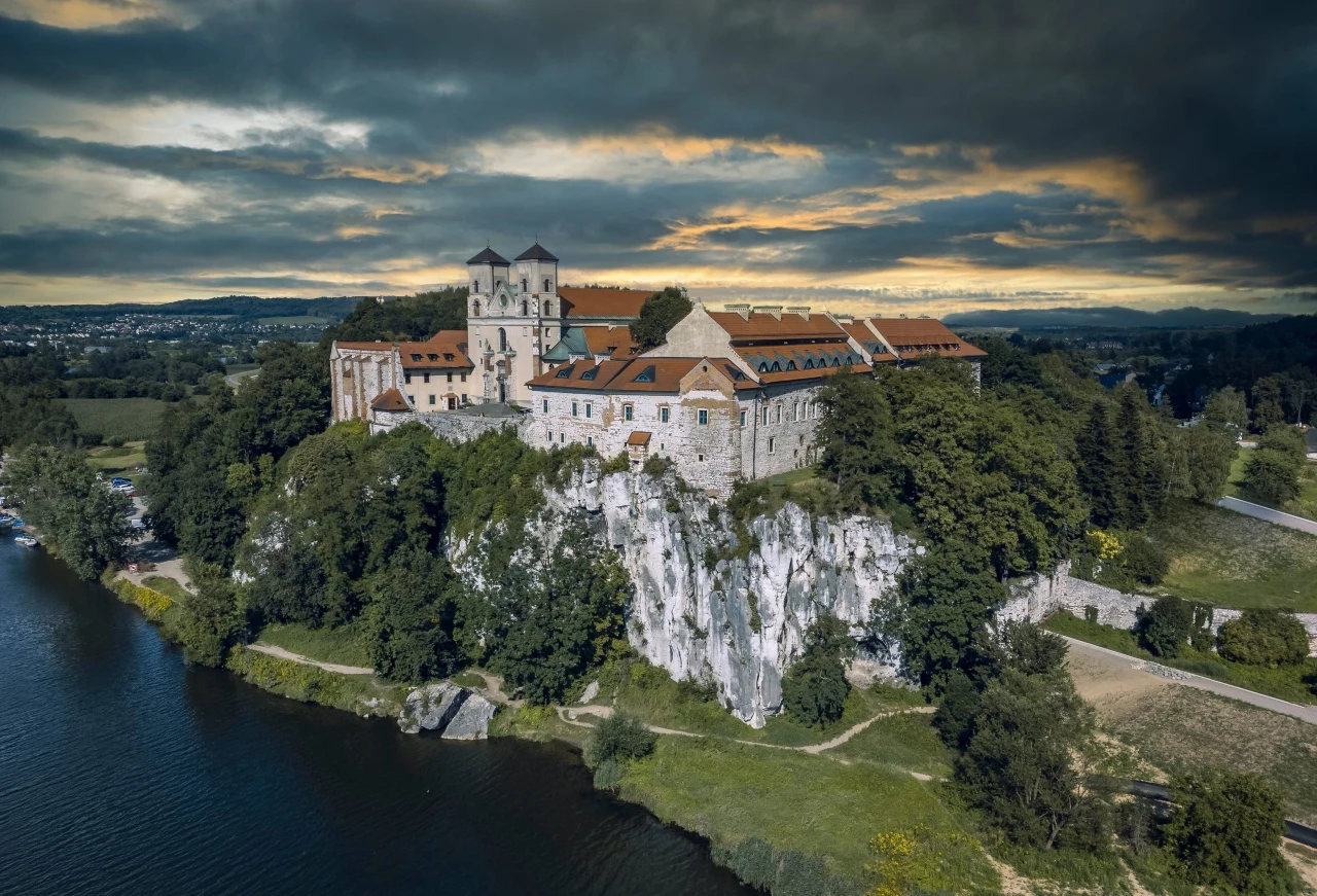 Opactwo benedyktynów w Tyńcu Opactwo benedyktynów w Tyńcu na wapiennej skale nad Wisłą, otoczone zielenią i polami, pod zachmurzonym niebem.