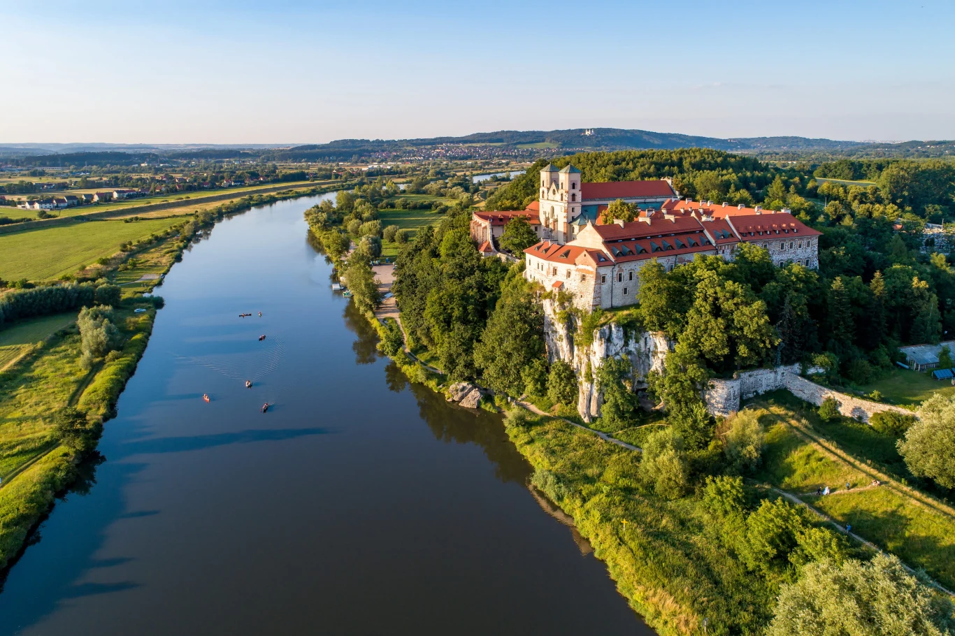 Opactwo w Tyńcu Skała nad szeroką rzeką z kompleksem klasztornym z czerwonymi dachami, otoczonym rozległą zielenią i łagodnym, letnim światłem.