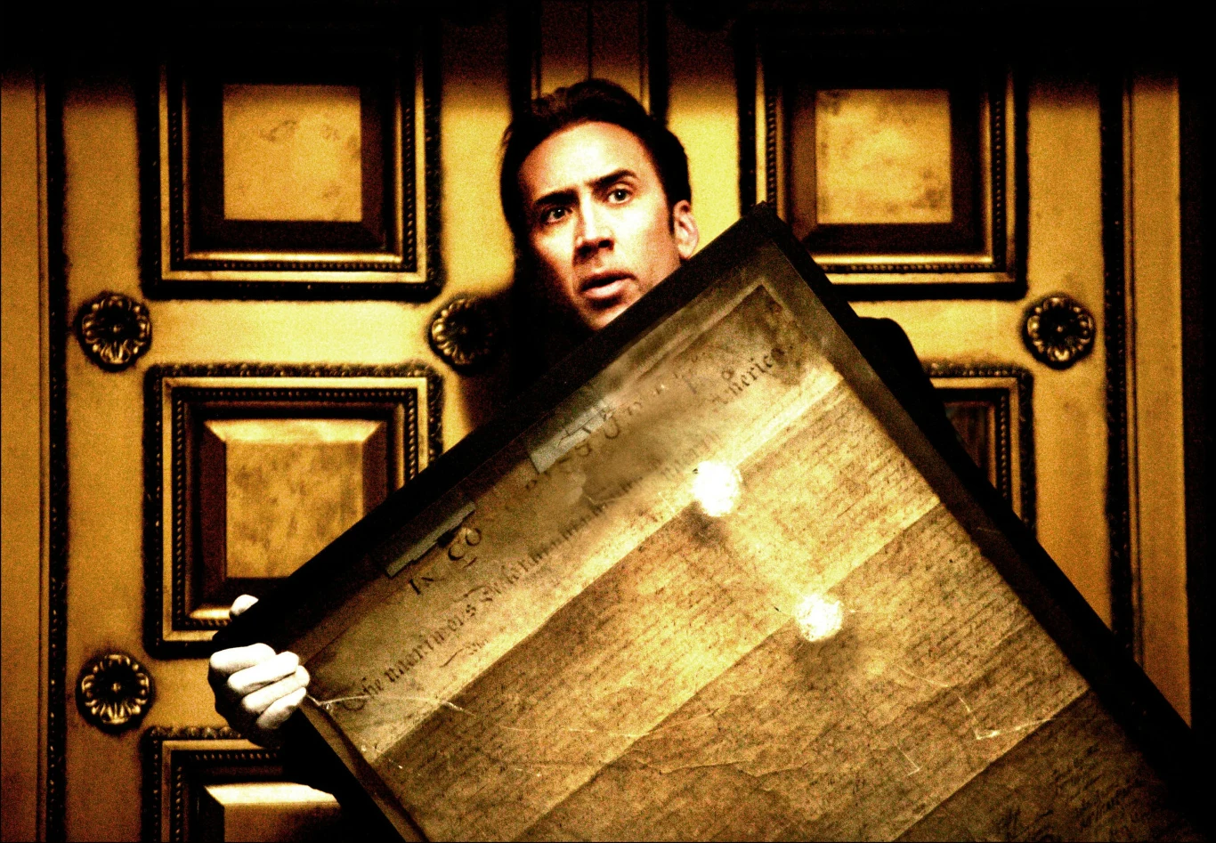 Nicolas Cage w filmie "Skarb narodów" Nicolas Cage trzyma starą kartę w ramie, stojąc pod złotymi drzwiami w filmie "Skarb narodów".