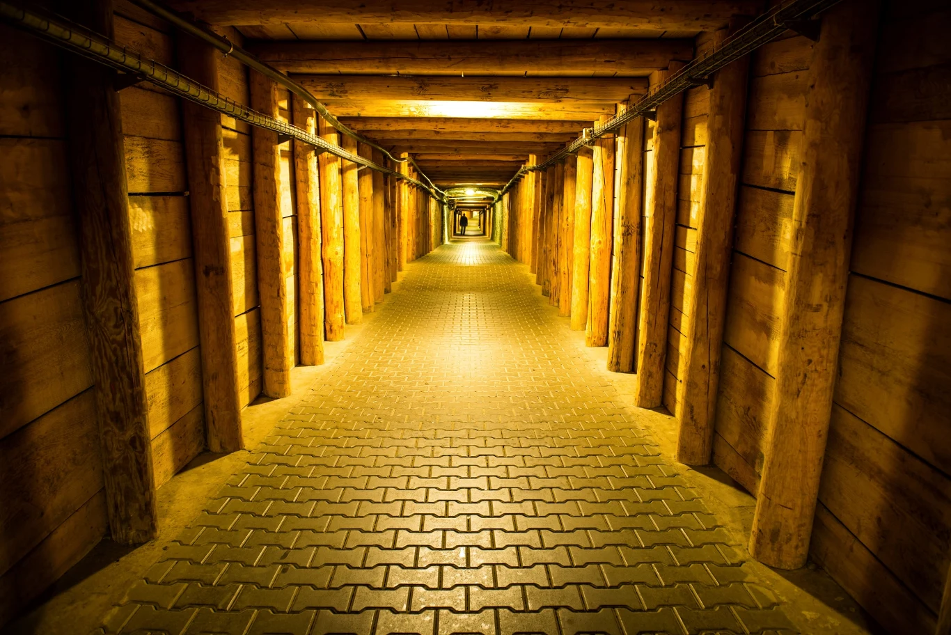 Drewniany tunel z żółtym oświetleniem, podparty grubymi belkami, prowadzący w głąb korytarza wyłożonego kostką brukową.