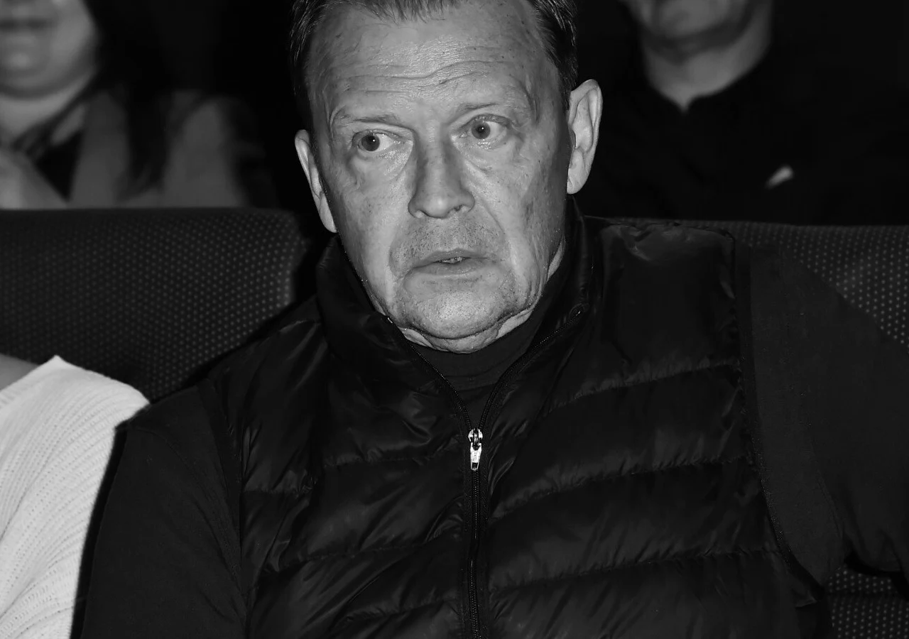 Jan Frycz Mężczyzna w średnim wieku w ciemnej, pikowanej kamizelce siedzi na widowni, wyraża zaskoczenie lub skupienie; w tle sylwetki innych osób.