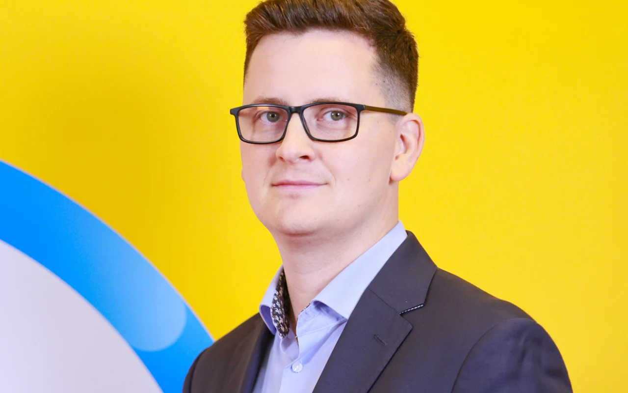 Rzecznik NFZ Łukasz Jarocki Mężczyzna w okularach, ubrany w granatową marynarkę i jasnoniebieską koszulę, stoi na tle intensywnie żółtej ściany z fragmentem niebiesko-białego okręgu po lewej stronie.