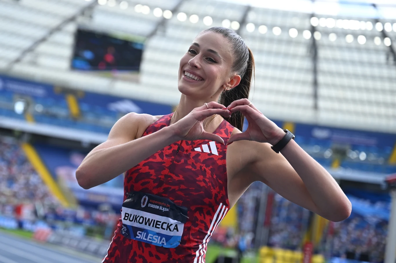 Uśmiechnięta lekkoatletka ubrana w czerwony strój sportowy z plakietką startową składa dłonie w kształt serca na tle stadionu lekkoatletycznego.