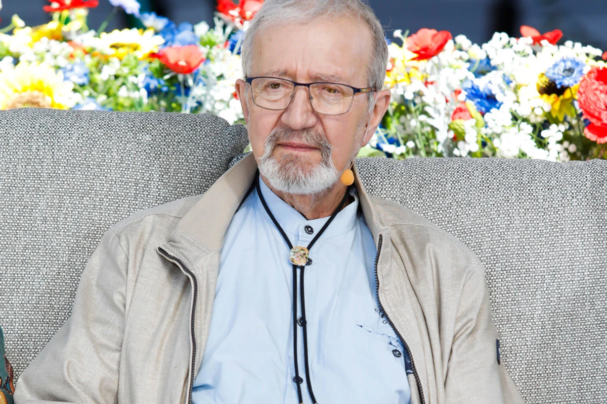 Krzysztof Daukszewicz Krzysztof Daukszewicz, starszy mężczyzna z siwą brodą i okularami, na szarej sofie, w jasnej kurtce, z mikrofonem przy twarzy