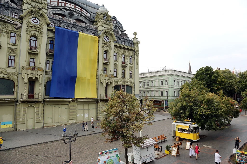 Ukraińskie władze ostrzegają przed wzmożonymi atakami Rosjan w Dzień Niepodległości Flaga Ukrainy na fasadzie zabytkowego budynku, plac z drzewami, żółty tramwaj, spacerujący ludzie