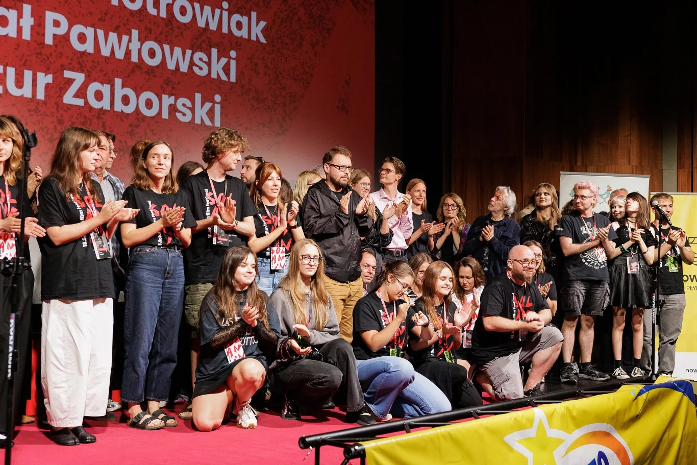 Gala finałowa 18. edycji Solanina Grupa osób stojących i siedzących na czerwonym dywanie, wielu z nich bije brawo, część trzyma identyfikatory na szyi, w tle widoczna czerwona ścianka z nazwiskami.