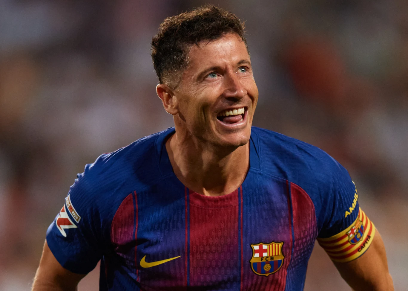 Piłkarz w koszulce FC Barcelony z opaską kapitana na ramieniu uśmiecha się szeroko podczas meczu.