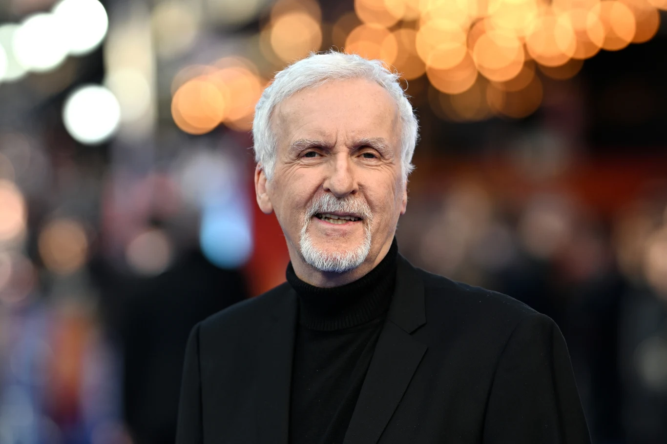 James Cameron Starszy mężczyzna z siwymi włosami i brodą, ubrany w ciemną marynarkę i golf, stoi na tle rozmazanych świateł, patrząc w stronę aparatu.