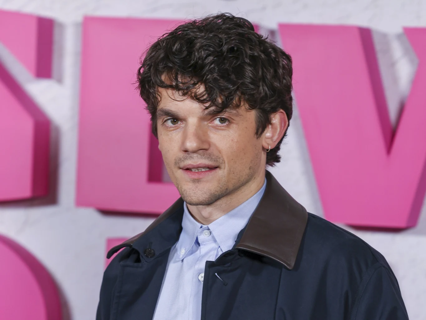 Edward Bluemel Mężczyzna o kręconych włosach ubrany w granatowy płaszcz i jasnoniebieską koszulę stoi na tle różowych, dużych liter.