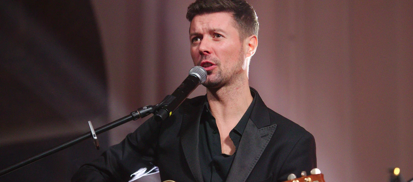 Maciej Szczepanik z "Pectus" Maciej Szczepanik w czarnej marynarce śpiewa do mikrofonu i gra na gitarze akustycznej na scenie.