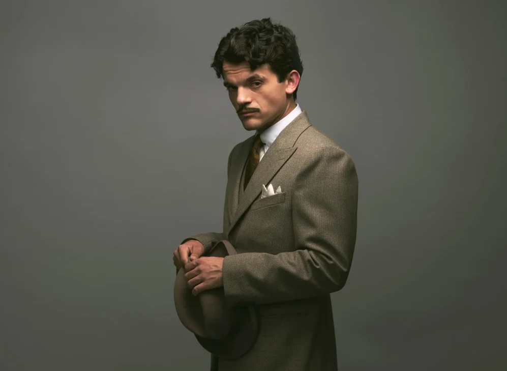 Edward Bluemel jako Hercules Poirot Mężczyzna w eleganckim, jasnobrązowym garniturze z białą poszetką i krawatem, trzymający kapelusz w dłoniach, patrzący poważnie w stronę aparatu na jednolitym, neutralnym tle.