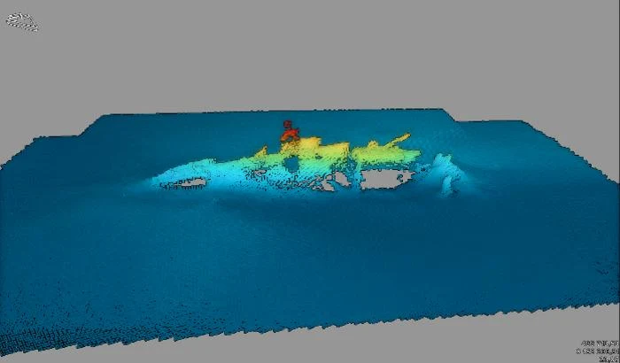 Model 3D wraku okrętu podwodnego UC-30, stworzony na podstawie skanów dna morskiego, uzyskanych za pomocą echosondy wielowiązkowej. Model rekina zanurzony w niebieskiej wodzie, z postacią w czerwonym stroju siedzącą na jego grzbiecie.