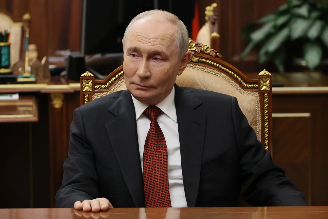 Rosja werbuje przestępców w Europie by atakować wspierające fabryki wspierające Ukrainę. Na zdj. Władimir Putin Władimir Putin w garniturze i czerwonym krawacie siedzi przy drewnianym biurku na rzeźbionym krześle.