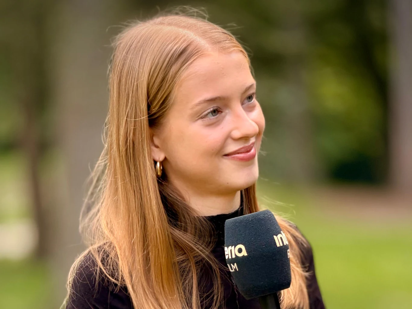 Karolina Ludwiniak Młoda kobieta z długimi blond włosami trzyma mikrofon z logo, uśmiechając się delikatnie do osoby poza kadrem, zieleń w tle
