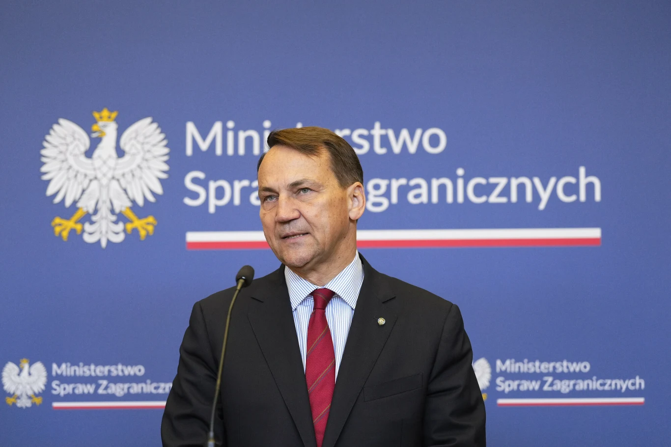 Radosław Sikorski Radosław Sikorski w garniturze i czerwonym krawacie przemawia na tle godła Polski i napisu Ministerstwo Spraw Zagranicznych.