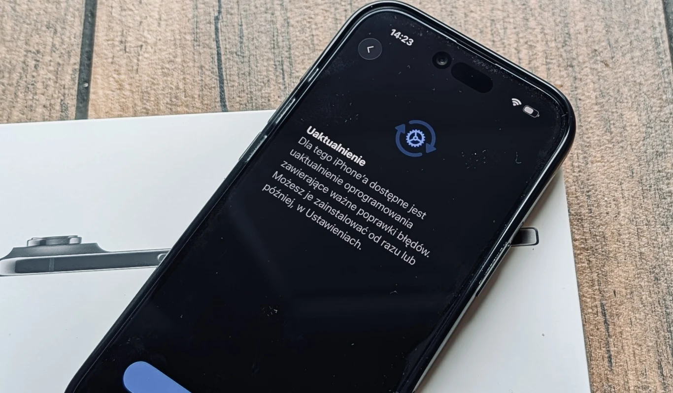 System iOS 27 zainstalowałem na iPhonie Air, gdy tylko stało się to możliwe. Czarny smartfon leżący na zamkniętym pudełku, z włączonym ekranem wyświetlającym komunikat dotyczący uaktualnienia systemu, na jasnym, drewnianym blacie.