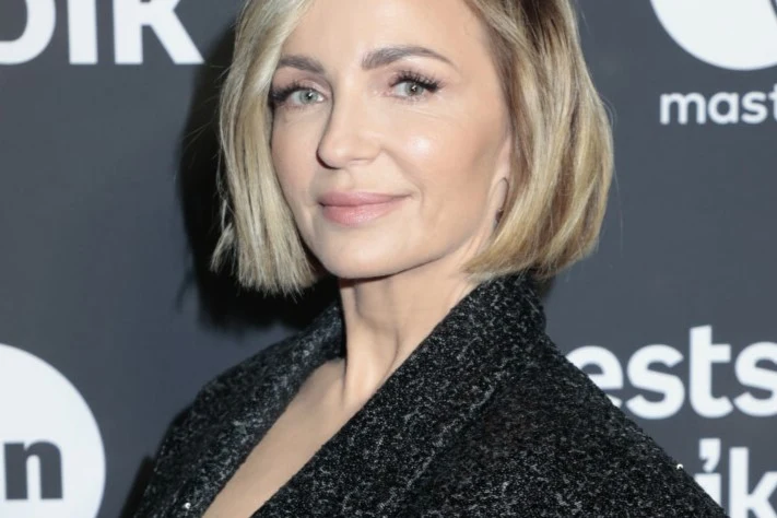 Ania Wyszkoni opublikowała nowe zdjęcia Ania Wyszkoni w krótkich blond włosach i czarnej stylizacji na tle ścianki z białymi napisami i logo.