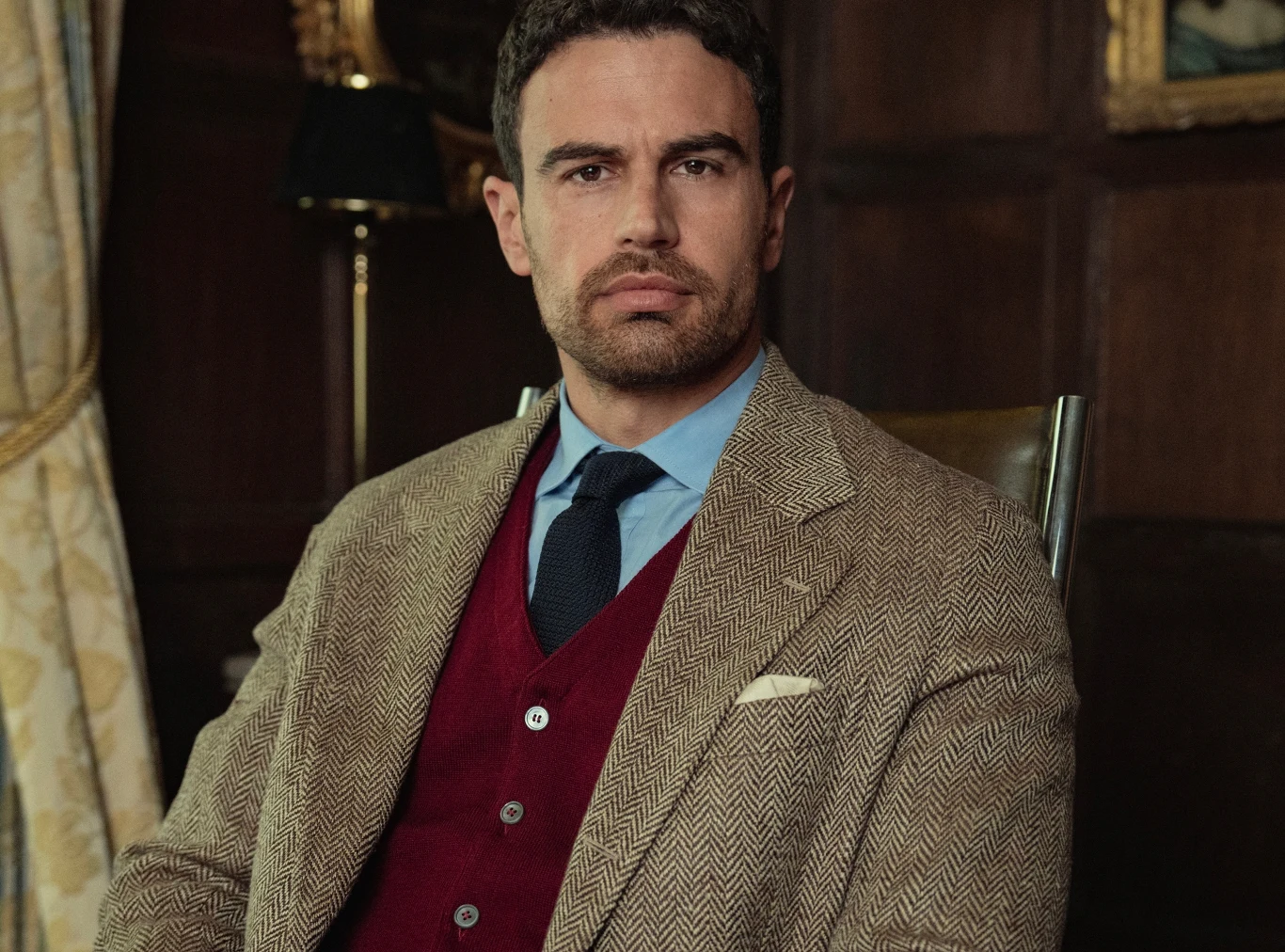 "Dżentelmeni" Theo James w beżowym garniturze i bordowym swetrze siedzi na tle ciemnego, drewnianego wnętrza.