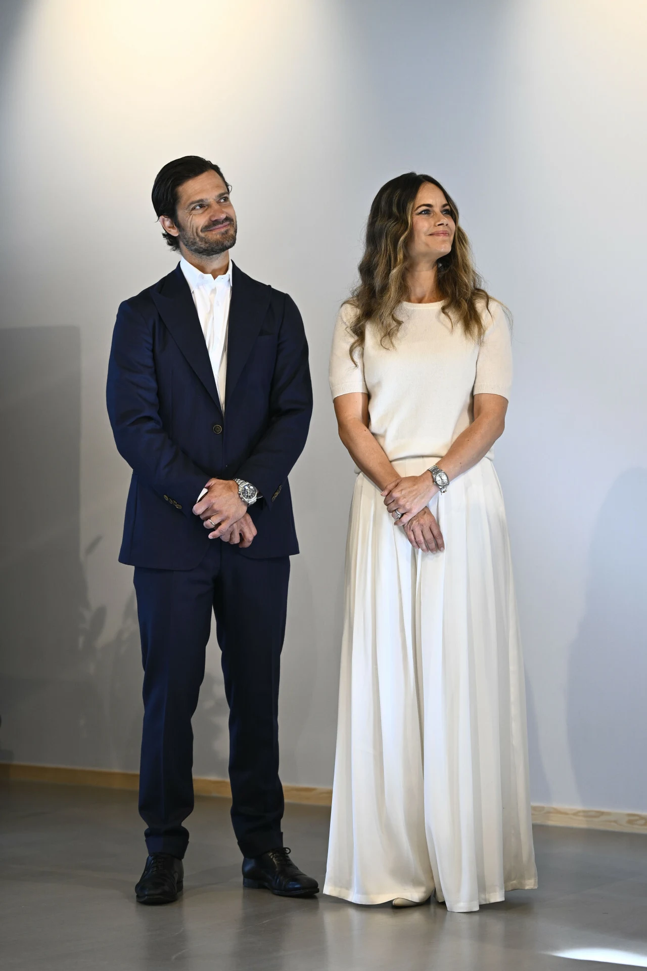 Elegancko ubrany mężczyzna w granatowym garniturze i kobieta w białej, długiej sukni stoją obok siebie, patrząc lekko w bok i uśmiechając się.