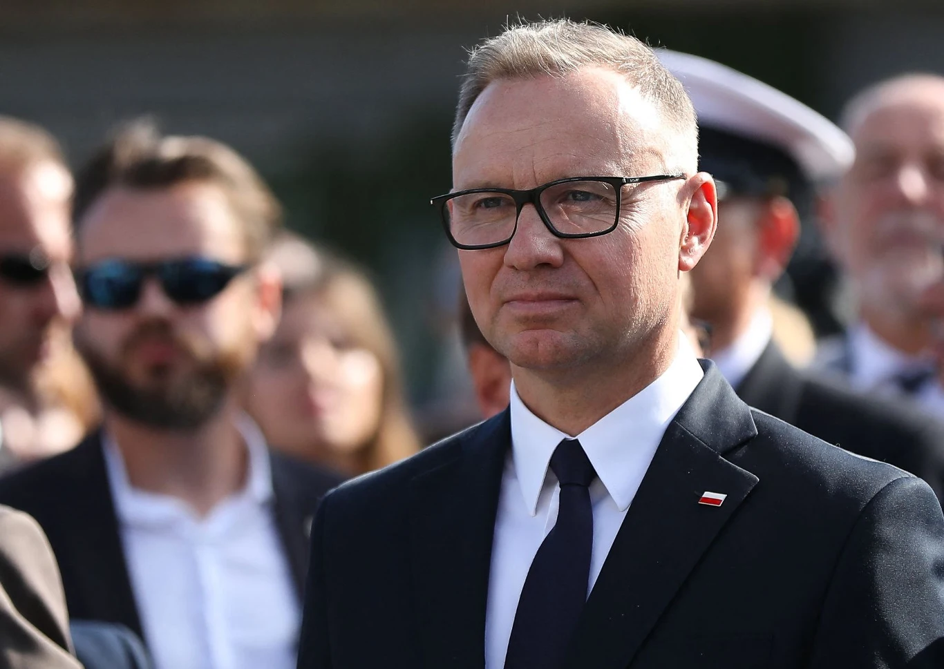 Andrzej Duda Mężczyzna w garniturze i okularach, z przypiętą do klapy marynarki polską flagą, stoi na tle grupy ludzi podczas oficjalnego wydarzenia.