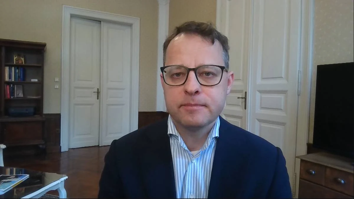 Marcin Romanowski Marcin Romanowski w okularach i granatowej marynarce, siedzi w eleganckim wnętrzu z białymi drzwiami.