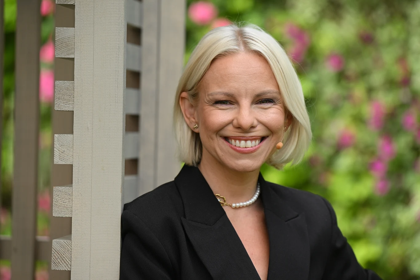 Alicja Janosz szykuje taneczną płytę Alicja Janosz, blond włosy, czarny żakiet, perłowy naszyjnik, stoi przy pergoli, w tle zieleń i różowe kwiaty.
